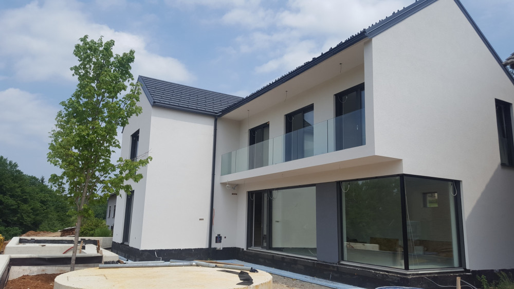 Moderna enodružinska hiša z belo fasado, dvokapno streho, velikimi steklenimi površinami in steklenim balkonom v naravnem okolju.