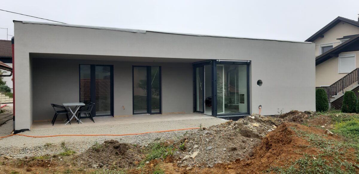 Moderna pritlična hiša z ravno streho, svetlo fasado, velikimi steklenimi površinami in pokrito teraso v fazi zaključevanja gradnje.