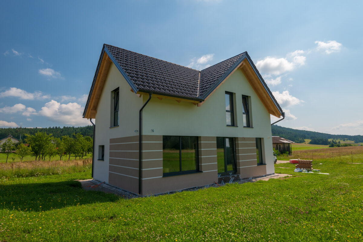 Novozgrajena enodružinska hiša na podeželju z moderno fasado v naravnih odtenkih, dvokapno streho, velikimi okni in zeleno travnato okolico v sončnem vremenu.