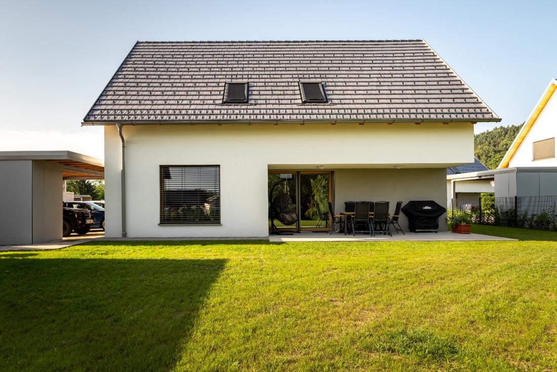 Sodobna družinska hiša s klasično dvokapno streho, svetlo fasado in prostorno pokrito teraso, obdana z urejenim zelenim vrtom.
